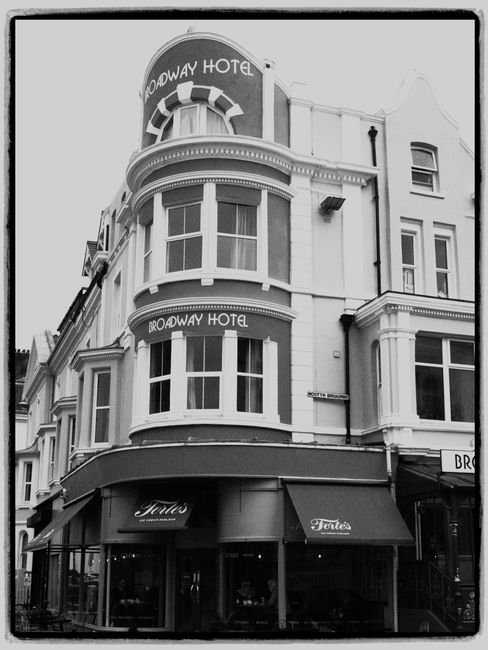 Black and white photograph of a multi-storey building with a curved façade, displaying signage for the Broadway Hotel at the top and middle levels. The ground floor features a storefront with large w