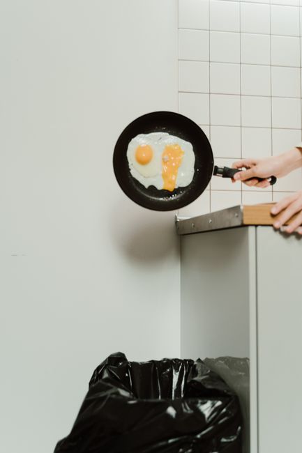 A person is holding a black frying pan with a wooden handle, in which a fried egg with a bright yellow yolk and cooked egg white is visible, positioned above a grey kitchen counter with a small overha