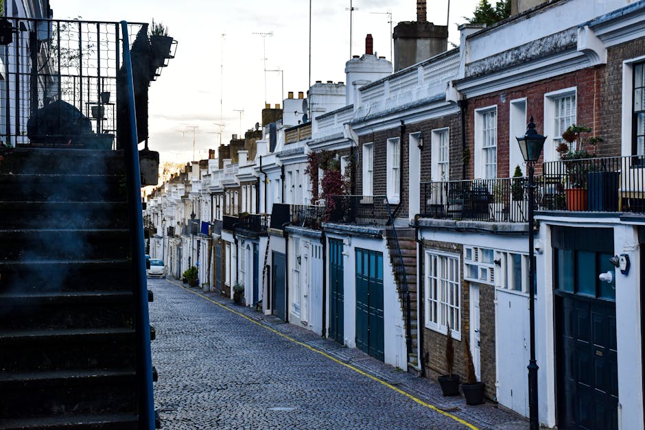 A street scene of a residential area during daytime with a row of terraced houses featuring brick and white facades, some with small front gardens and potted plants on the balconies. On the left side,