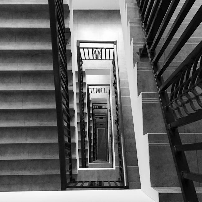 A narrow, spiral staircase with metal steps and a black handrail ascending in a confined indoor space, adjacent to a white wall with visible piping and a window at the top. The blue painted steps at t